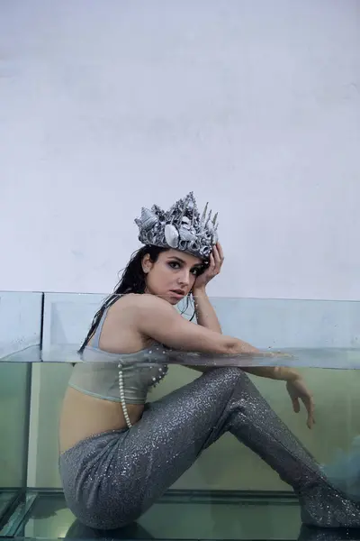 stock image A young woman, dressed as a mermaid, sits in a glass tank, wearing a crown of shells and pearls.