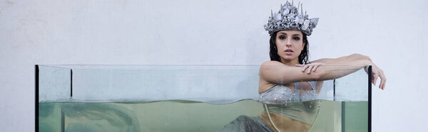A young woman dressed as a mermaid leans against the edge of an aquarium, wearing a silver crown.