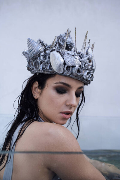 A young woman with long wet hair and a silver seashell crown looks down at the water, submerged in a tank.