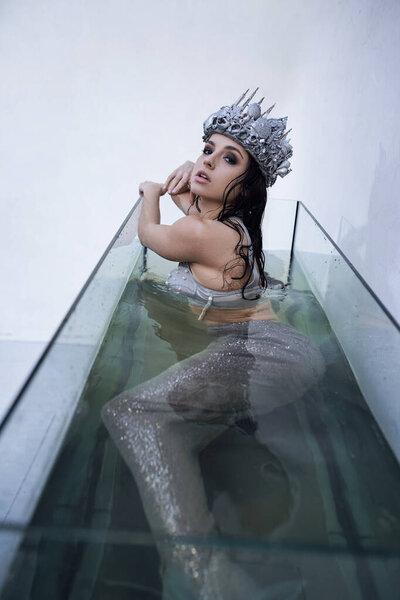 A young woman, dressed as a mermaid, with long dark hair, sits in a glass enclosure filled with water, wearing a silver crown.