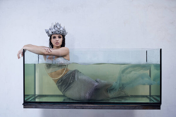 Молодая женщина, украшенная блестящей короной и хвостом русалки, смотрит изнутри наполненного водой бака.