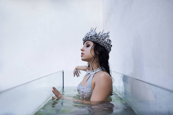 Молодая женщина в короне, украшенная серебром, плавает в современном, стеклянном аквариуме.