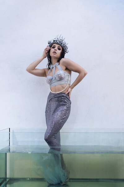 A young woman in a mermaid costume stands in a glass tank filled with water, her tail shimmering.