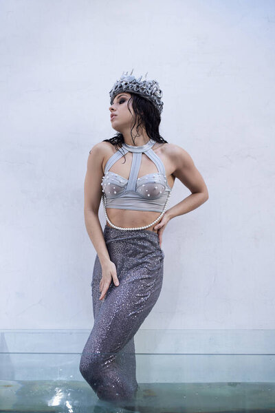 A young woman, adorned in a silver crown and a shimmering tail, stands in a pool of water, her gaze fixed on the horizon.
