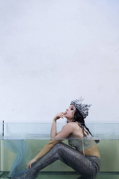 A young woman, adorned in a crown, sits in a tank of water, resembling a mermaid.