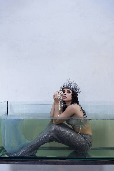 A mermaid sits in a tank of water, wearing a crown and holding a string of pearls.