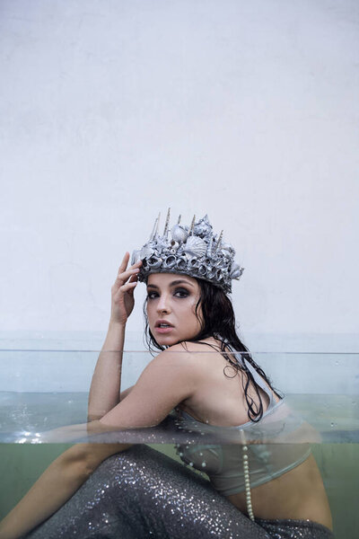 A young woman wearing a silver seashell crown sits in a glass tank filled with water, her mermaid tail shimmering beneath the surface.