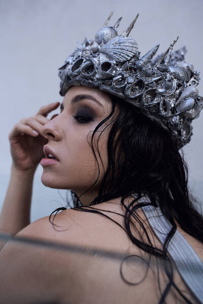 A young woman with dark hair wears a crown of seashells while immersed in water, her face gazing dreamily off to the side.