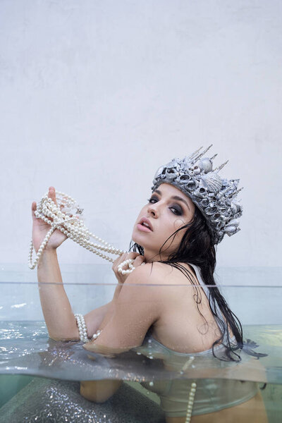 A young woman dressed as a mermaid with a seashell crown sits in a water tank, holding a pearl necklace.