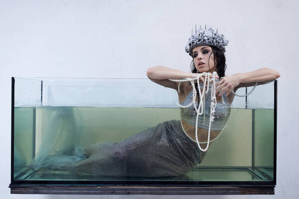 A young woman, dressed as a mermaid, sits in a glass tank, holding a string of pearls.