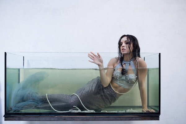 A young woman, dressed as a mermaid, floats in a glass tank, her eyes gazing upward with a look of longing.
