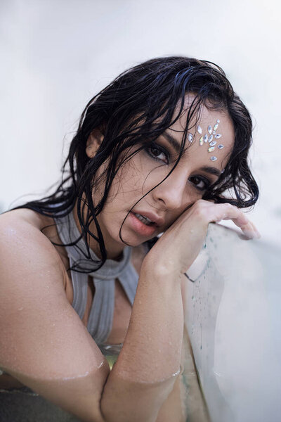 A young woman with dark hair and rhinestones adorning her forehead rests her head against the wall of an aquarium.