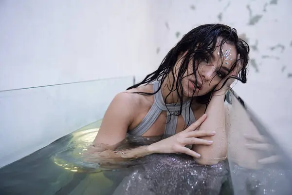 stock image A young woman with dark hair and a sparkling gaze rests her head on the edge of an aquarium, her body submerged in the water.