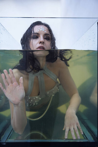 Dark-haired woman in shimmering top emerges from water in glass tank, looking sad yet hopeful.