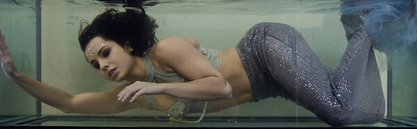 A young woman with long brunette hair swims in a large aquarium, dressed as a mermaid.