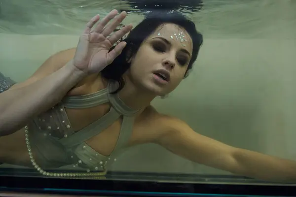 stock image A young woman, adorned with shimmering makeup, swims in an aquarium, her eyes fixed on the viewer.