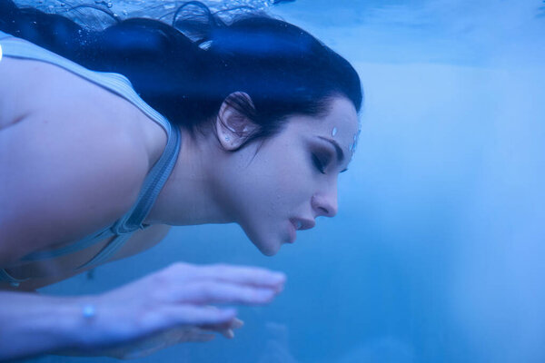 A young woman with dark hair, dressed in a white top, swims gracefully underwater in an aquarium.