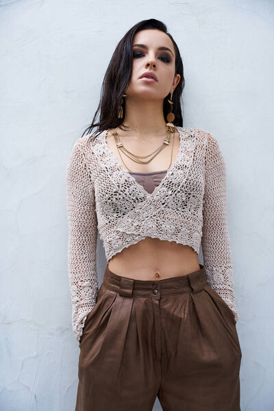 A young woman with long brunette hair poses in a white knitted top and brown leather pants against a white backdrop.