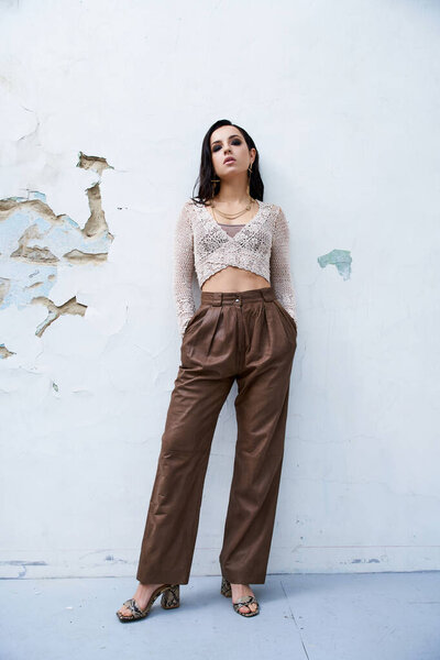 A young woman with long brunette hair poses in a white knit top and brown pants against a white wall.