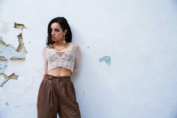 A young woman with long, dark hair wears a white knitted top and poses against a studio backdrop.