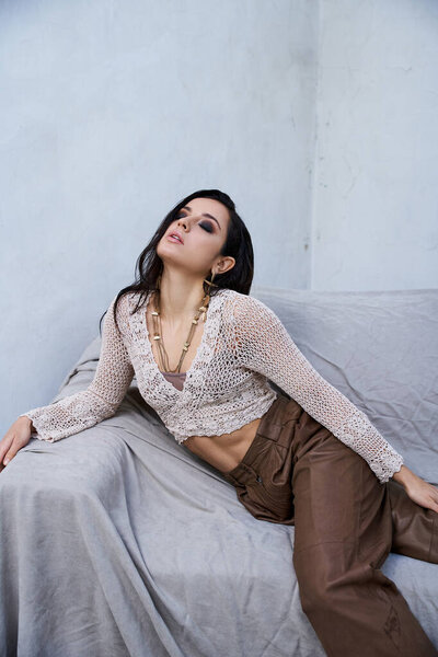 A young woman with long, dark hair wears a white knit top and brown pants while posing on a gray couch in a studio setting.