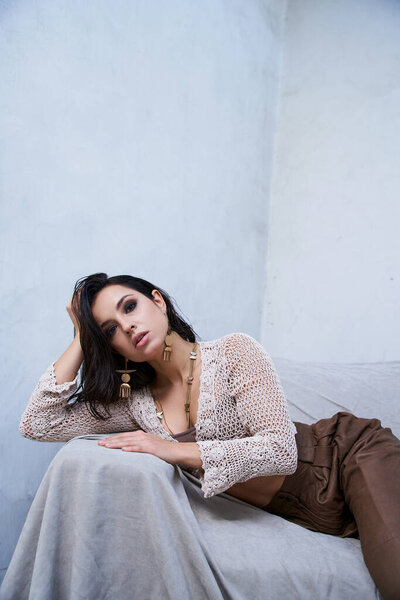 A tanned brunette model poses in a white knitted top and brown pants against a white backdrop.