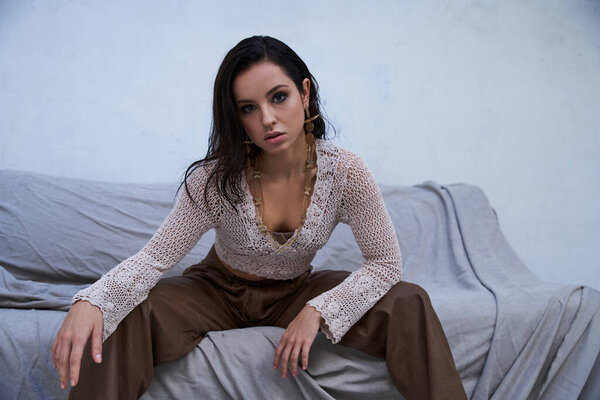 A young woman with long brunette hair and a tanned body sits on a couch, wearing a white knit top and brown pants.
