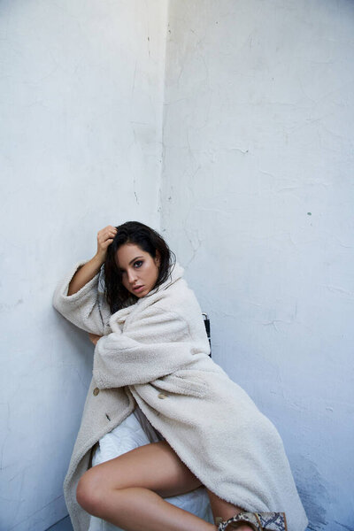 A young woman with long brunette hair poses in a white teddy coat in a studio setting.