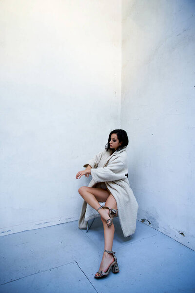 A young woman with brunette hair poses in a white teddy coat, sitting on a chair in a studio setting.
