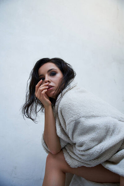 A young woman in a white teddy coat sits against a white wall, gazing softly.