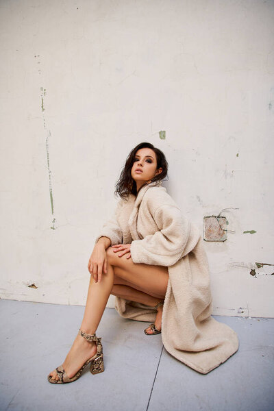 A brunette woman poses in a white teddy coat and snakeskin heels against a plain white background.