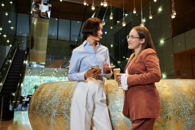 Modern bir ofis lobisinde iki meslektaş sohbet edip kahveyi paylaşıyor..