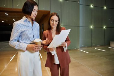 İki iş kadını modern bir ofis lobisinde durmuş kahve fincanlarını tutarken evrak işlerini tartışıyorlar..