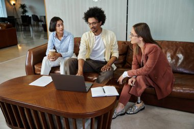 Üç meslektaş modern bir ofis lobisinde birlikte oturup bir dizüstü bilgisayar ve diğer belgeleri tartışıyorlar..