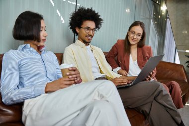 Three colleagues sit on a couch in an office lobby, chatting and looking at a laptop. clipart