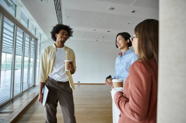 Üç meslektaş modern bir ofiste durmuş, sohbet ediyor ve kahve fincanları tutuyorlar..