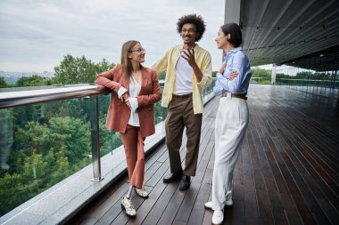 Three colleagues chat on a modern rooftop terrace with a view of trees and cityscape. clipart