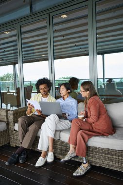 Diverse colleagues relax on a rooftop terrace, enjoying a casual work conversation. clipart