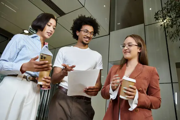 stock image Three colleagues, engage in conversation while holding coffee and documents in an office lobby.