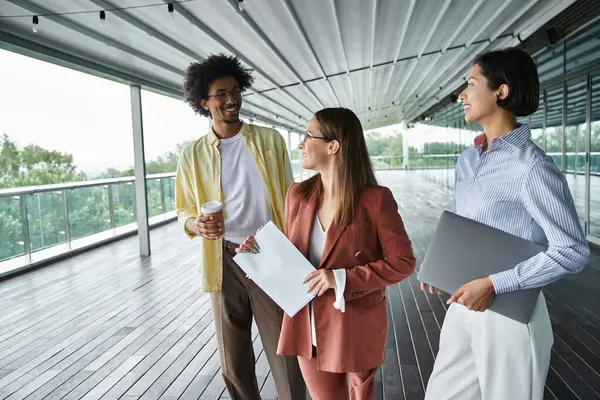 Stock image Three diverse colleagues chat and collaborate on a modern office rooftop terrace.