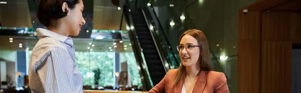 stock image Two coworkers chat and smile in an office lobby. One wears a striped shirt, the other a blazer and glasses.