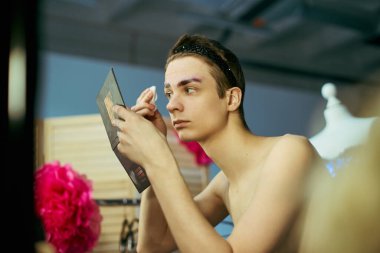 A drag queen prepares for their performance by applying makeup in front of a mirror backstage. clipart