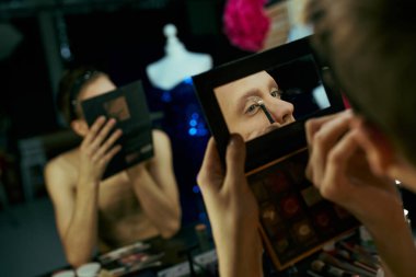 Bir travesti sahne arkasına göz kalemi sürüyor, aynadaki yansımasına odaklanıyor..
