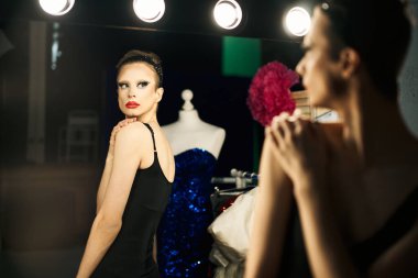 Siyah elbiseli bir travesti, renkli bir ışık ve elbise formuyla çevrili bir aynanın önünde gösteriye hazırlanıyor.