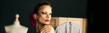 Makyajlı ve saç bandı takmış bir travesti, makyaj masasının yanına oturur ve gösteriye hazırlanır..