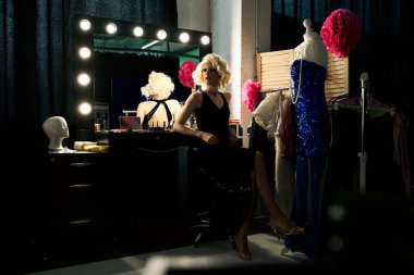 Bir travesti soyunma masasında oturuyor, bir gösteri için hazırlanıyor, yanında bir manken kostümü var..