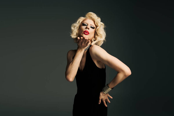 A drag queen in a black dress and blonde wig poses against a grey background, blowing a kiss to the camera.