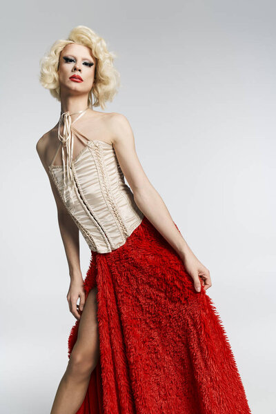 Королева трансвеститов в светлом парике, красной юбке и корсете позирует на сером фоне.