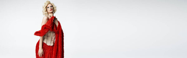Королева трансвеститов в светлом парике и драматических позах в красной юбке и корсете на сером фоне.