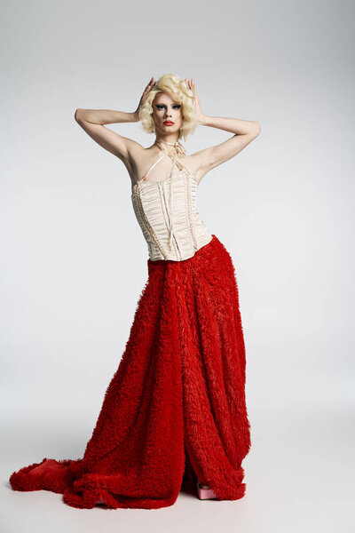 Королева трансвеститов в светлом парике и смелых позах макияжа в красной юбке и белом корсете на сером фоне.
